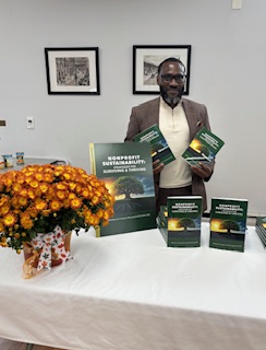 Ajené smiling behind a table of copies of Nonprofit Sustainability, holding the book up beside a display of the cover.