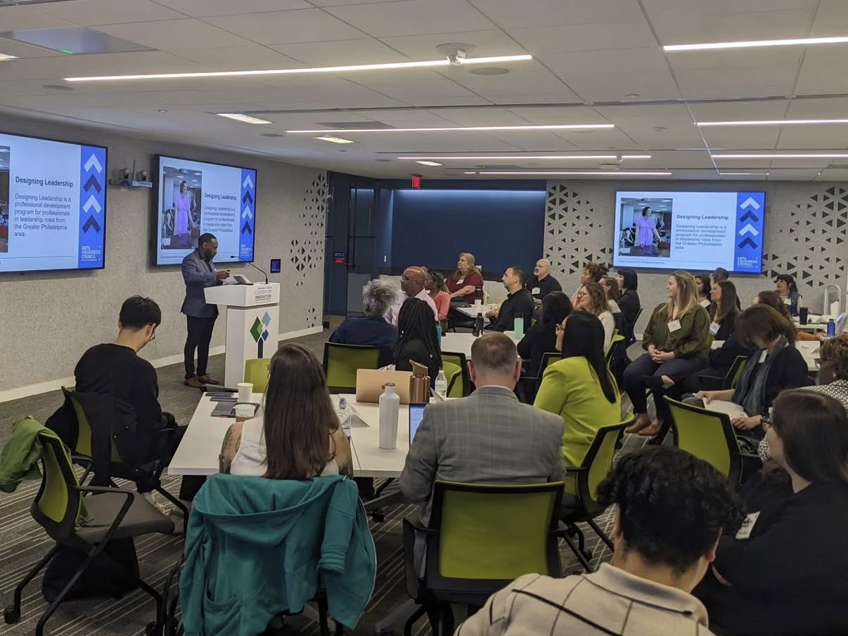 Ajené presenting at the lectern to a full room of professionals during an Arts + Business Council Designing Leadership session, program slides on the screens.